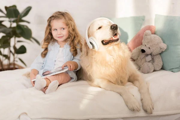 Adorable kid holding tablet, funny golden retriever with headphones lying on bed in children room — Stock Photo