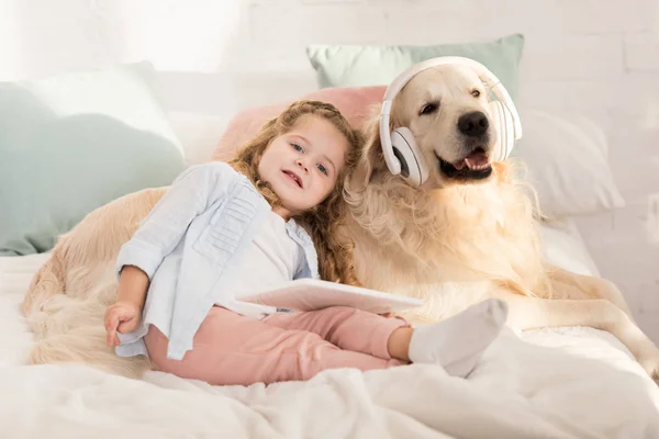 Criança adorável segurando tablet e inclinando-se em bonito golden retriever com fones de ouvido deitado na cama no quarto das crianças — Fotografia de Stock