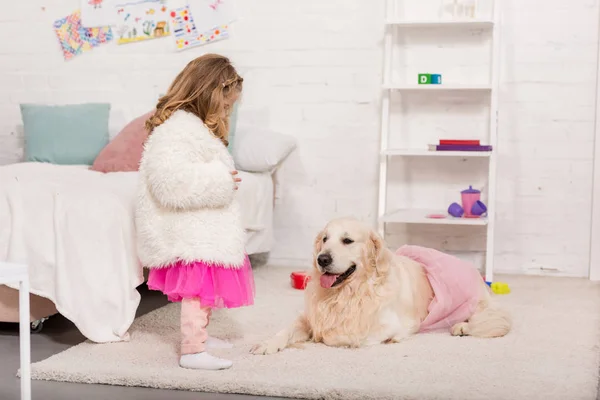 Kid in skirt looking at golden retriever lying on carpet in pink skirt in children room — Stock Photo