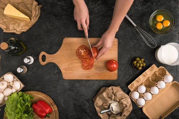 Vista cortada de mulher cortando tomates na tábua de corte com ingredientes de pizza em fundo cinza — Fotografia de Stock