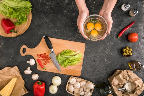 Partial view of woman holding bowl with eggs near pizza ingredients on grey background — Stock Photo