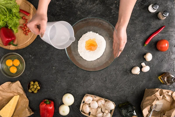 Vue partielle de la femme versant de l'eau dans un bol de farine avec des ingrédients de pizza sur fond gris — Photo de stock