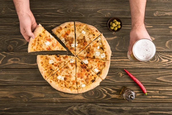 Vista ritagliata dell'uomo che mangia pizza al formaggio sul tavolo di legno con birra e olive — Foto stock