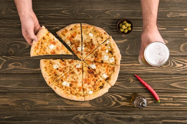 Cropped view of man eating cheese pizza on wooden table with beer and olives — Stock Photo