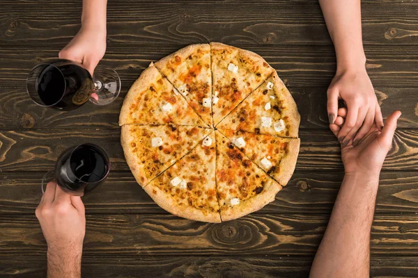 Vue recadrée du couple tenant les mains et verres de vin rouge tout en mangeant de la pizza sur une table en bois — Photo de stock