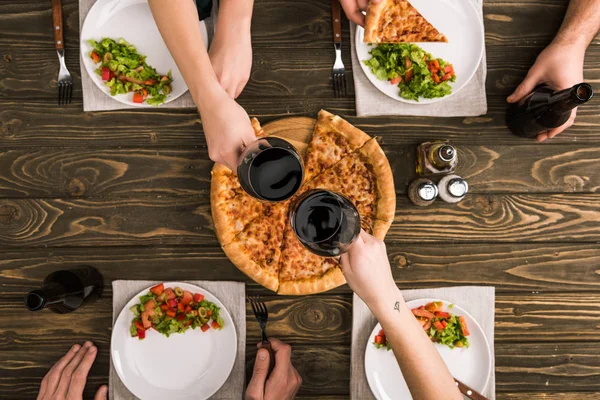 Vista parcial de amigos brindando mientras cenan con pizza y ensaladas en la mesa de madera - foto de stock