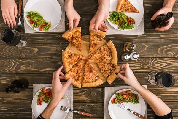 Обрезанный вид друзей, разделяющих пиццу за ужином с салатами за деревянным столом — стоковое фото