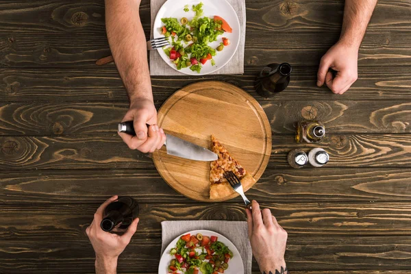 Abgeschnittene Ansicht von Männern, die sich Pizzascheiben mit Messer und Gabel am Holztisch teilen — Stockfoto