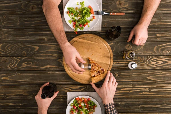 Vista parcial de los hombres condimento rebanada de pizza durante la comida en la mesa de madera - foto de stock