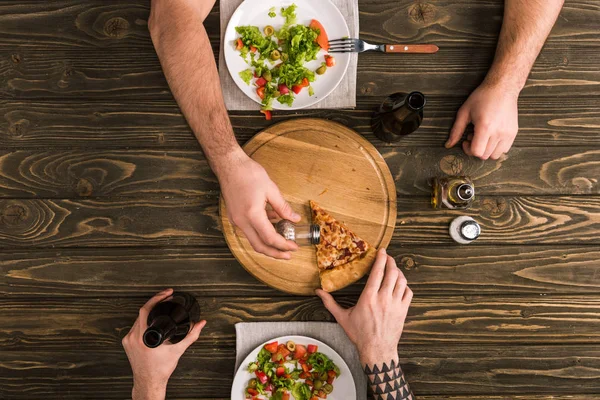 Vista parcial de los hombres condimento rebanada de pizza durante la comida en la mesa de madera — Stock Photo