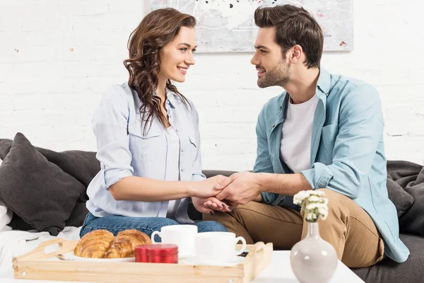 Smiling couple sitting on couch, looking at each other and holding hands while having breakfast at home — Stock Photo