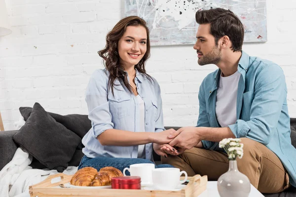 Coppia seduta sul divano e che si tiene per mano mentre fa colazione a casa — Foto stock