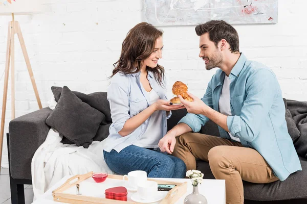Sonriente pareja comiendo croissants durante el desayuno en casa - foto de stock