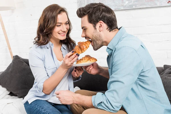 Schöne lächelnde Frau füttert Mann mit Croissant beim Frühstück zu Hause — Stockfoto