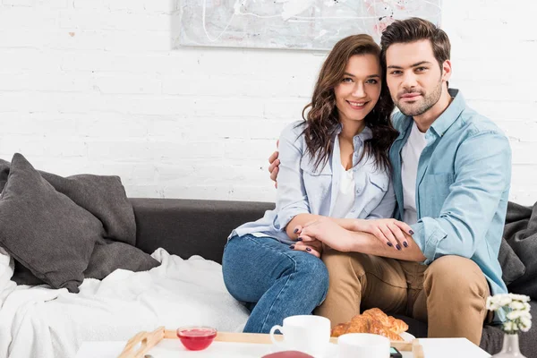 Pareja sentada en el sofá, mirando a la cámara y abrazando mientras desayuna en casa - foto de stock