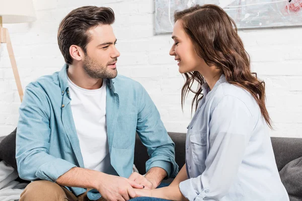 Couple sitting on couch, looking at each other and holding hands at home — Stock Photo