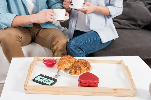 Vista cortada do casal sentado no sofá e beber café durante o café da manhã em casa — Fotografia de Stock