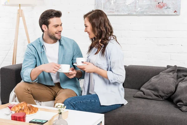 Couple souriant assis sur le canapé et boire du café pendant le petit déjeuner à la maison — Photo de stock