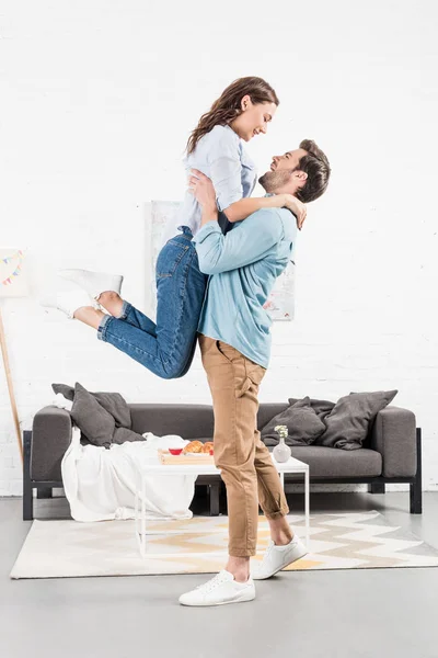 Hombre feliz en ropa casual sosteniendo mujer en la sala de estar - foto de stock