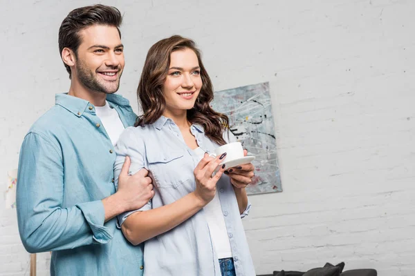 Smiling man tenderly embracing beautiful woman holding cup of coffee at home — Stock Photo