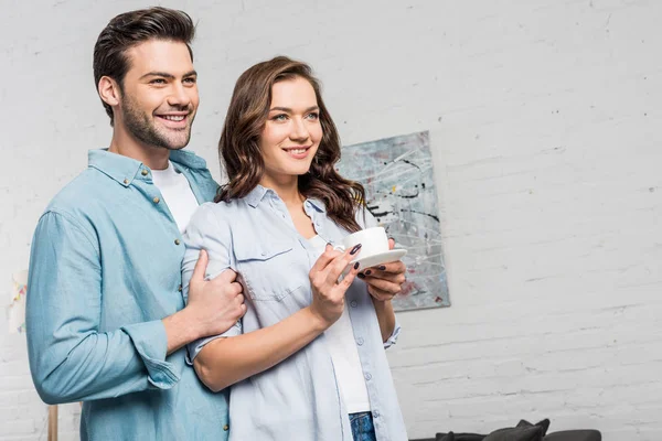 Sonriente hombre tiernamente abrazando hermosa mujer sosteniendo taza de café en casa - foto de stock