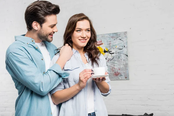 Hombre tiernamente abrazando hermosa mujer sonriente sosteniendo taza de café en casa - foto de stock