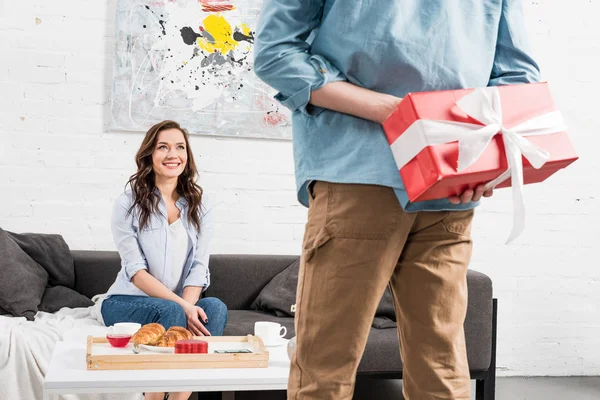 Vista posteriore di uomo in possesso di regalo di compleanno rosso dietro la schiena con donna sorridente sullo sfondo — Foto stock