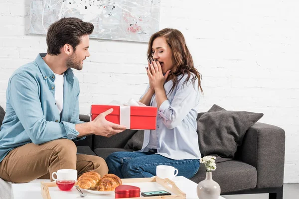 Hombre sentado en el sofá y la presentación de caja de regalo de cumpleaños a la mujer sorprendida en la sala de estar - foto de stock