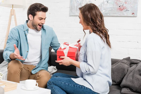 Mujer sentada en el sofá y presentando la caja de regalo de cumpleaños al hombre sorprendido en la sala de estar — Stock Photo