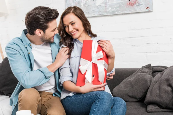 Bella coppia felice seduta sul divano, abbracciare e tenere regalo di compleanno in soggiorno — Foto stock
