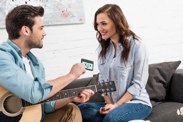 Mann mit Akustikgitarre sitzt auf Couch und gibt schöne Frau zu Hause Vintage-Audio-Kassette — Stockfoto