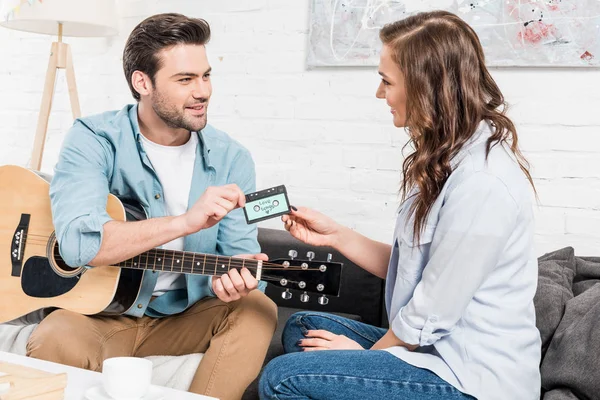 Handsome man with acoustic guitar sitting on couch and giving vintage audio cassette to woman at home — Stock Photo