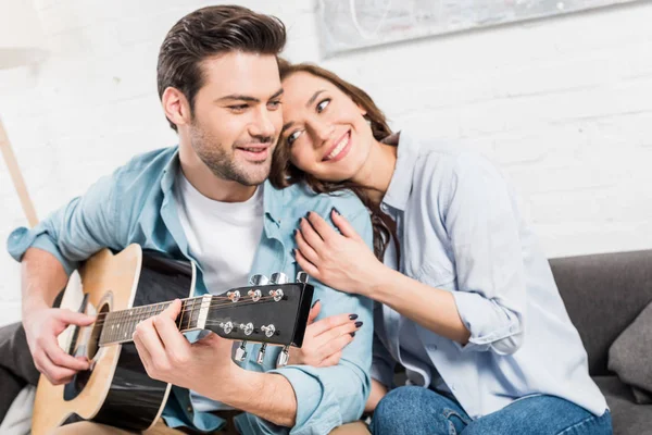 Coppia seduta e abbracciata sul divano mentre l'uomo suona la chitarra acustica a casa — Foto stock