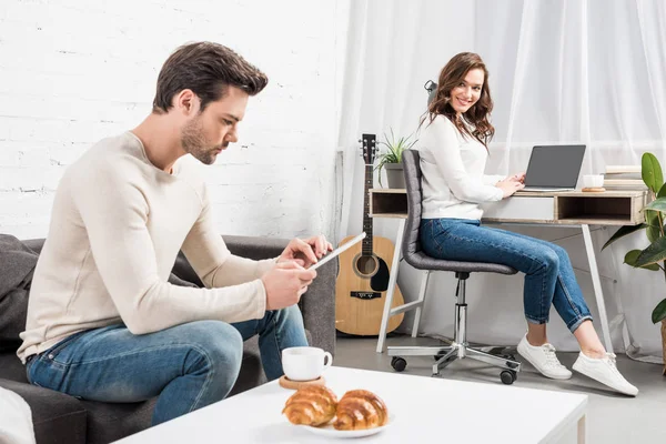 Hombre usando tableta digital mientras sonríe mujer sentada en el escritorio de la computadora en el fondo en la sala de estar - foto de stock