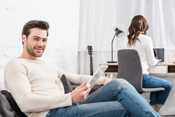 Улыбающийся мужчина с помощью цифрового планшета в то время как женщина сидит за столом компьютера на заднем плане в гостиной — стоковое фото