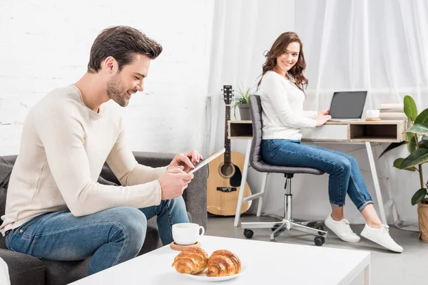 Homme utilisant tablette numérique tandis que belle femme assise au bureau de l'ordinateur sur fond dans le salon — Photo de stock