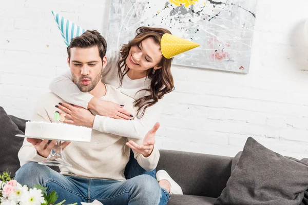 Happy couple in party hats hugging and celebrating birthday with cake at home — Stock Photo