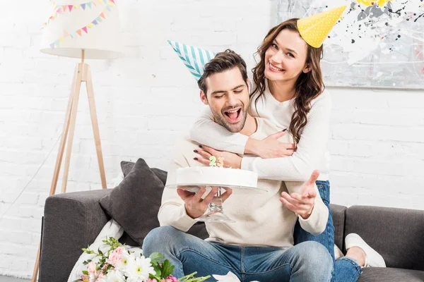 Couple gai chapeaux de fête étreignant et célébrant anniversaire avec gâteau à la maison — Photo de stock