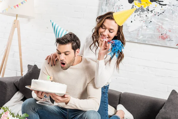Hombre soplando velas en pastel de cumpleaños mientras mujer animando con cuerno de fiesta en casa — Stock Photo