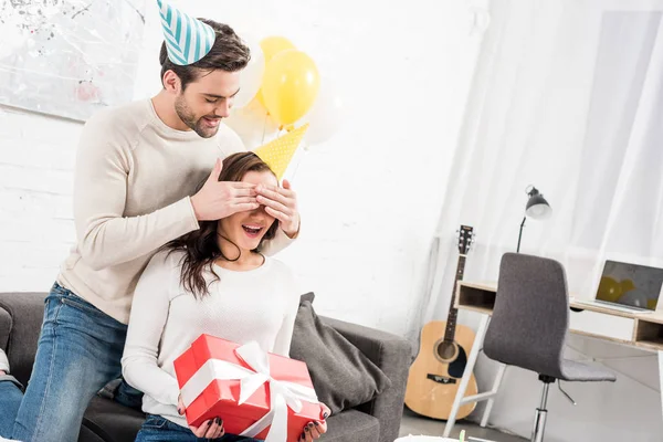 Homme en chapeau de fête fermer les yeux et faire surprise d'anniversaire avec cadeau pour femme excitée à la maison — Photo de stock