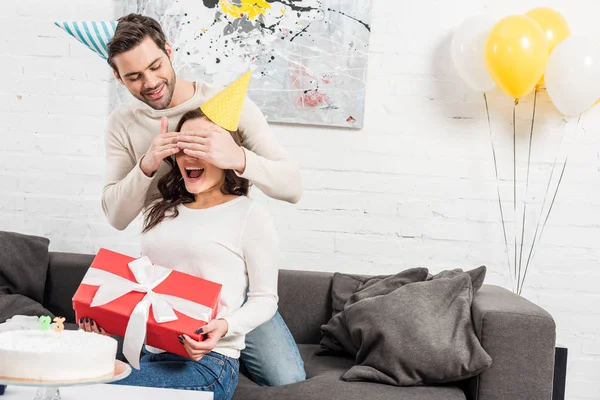 Mann überrascht aufgeregte Frau mit Geburtstagsgeschenk im Wohnzimmer — Stockfoto