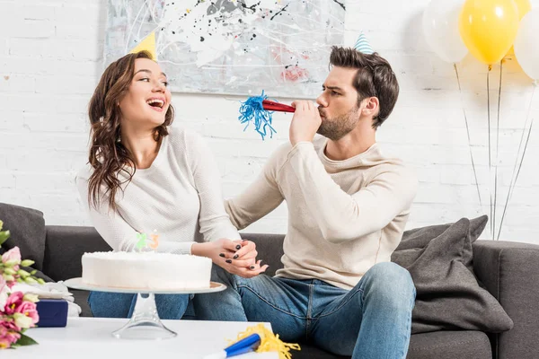 Paar in Partyhüten feiert Geburtstag mit Kuchen, während Mann zu Hause in Partyhorn bläst — Stockfoto