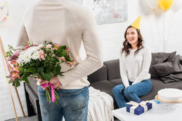 Vista trasera del hombre sosteniendo flores de cumpleaños detrás de la espalda con la mujer sentada en el sofá - foto de stock