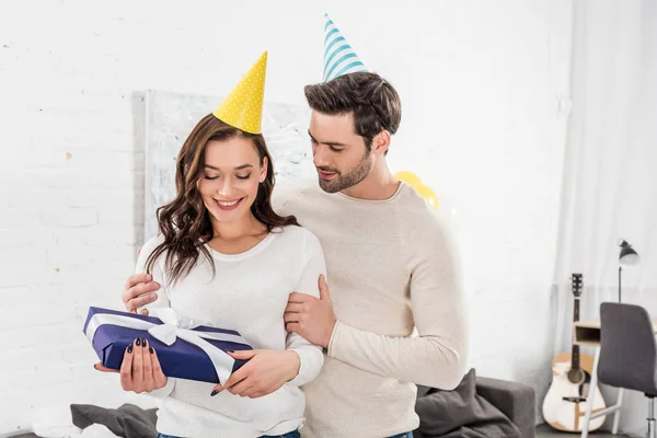 Lächelndes Paar mit Party-Hüten, das sich umarmt, während es Geburtstagsgeschenk im Wohnzimmer hält — Stockfoto