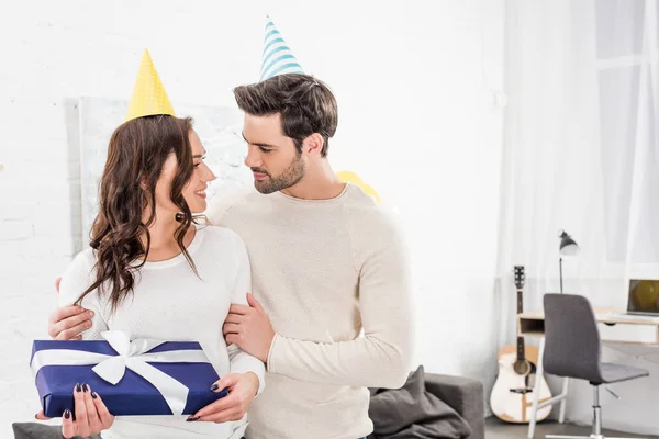Hermosa pareja en sombreros de fiesta abrazando mientras sostiene regalo de cumpleaños en la sala de estar - foto de stock