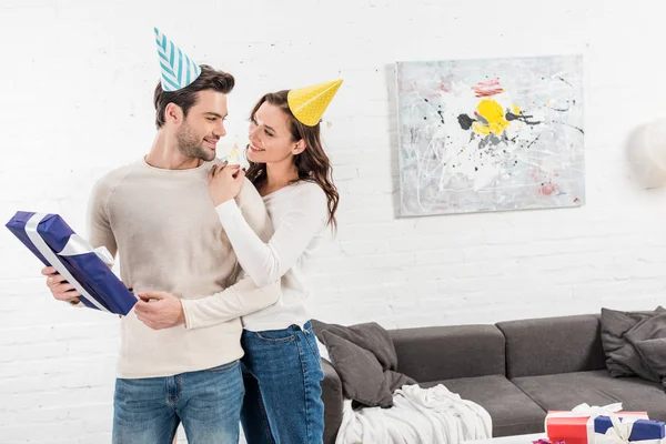 Hermosa pareja sonriente con caja de regalo mirándose, abrazando y celebrando cumpleaños en la sala de estar - foto de stock