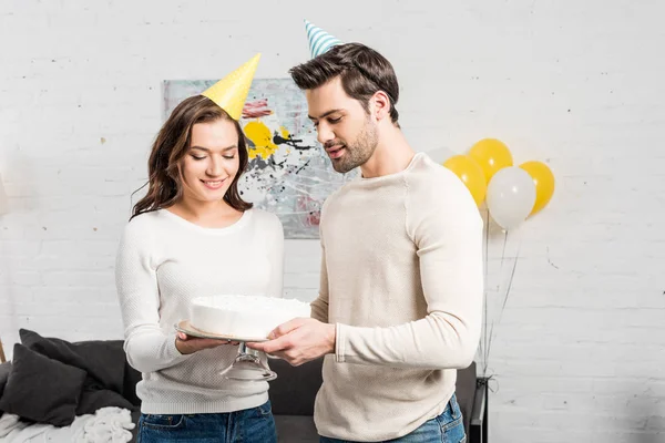 Smiling couple in party hats holding cake and celebrating birthday in living room — Stock Photo