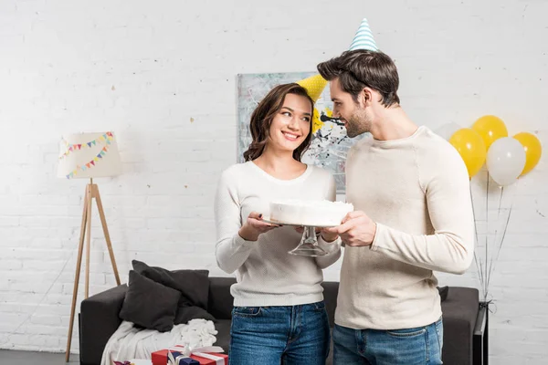 Feliz pareja en sombreros de fiesta celebración de pastel y la celebración de cumpleaños en la sala de estar — Stock Photo