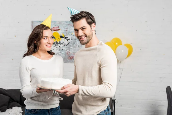 Belo casal em chapéus de festa segurando bolo e celebrando aniversário na sala de estar — Fotografia de Stock