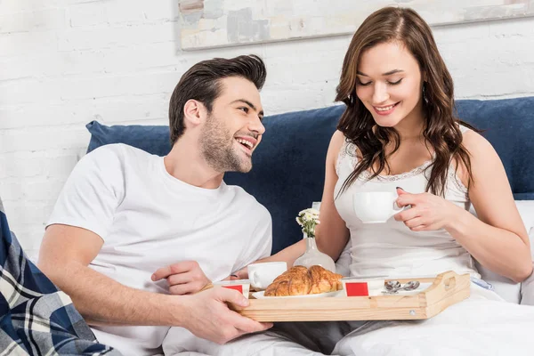 Happy couple lying in bed and having breakfast in morning at home — Stock Photo